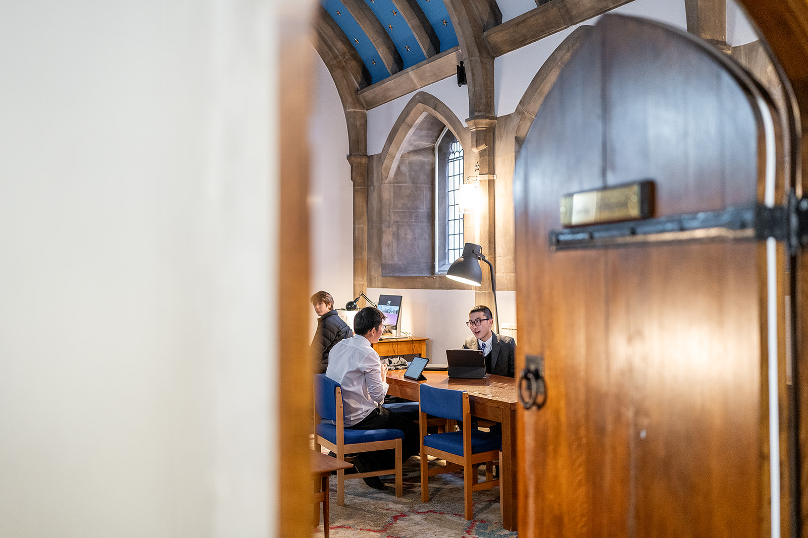 Through a door into a Common Room in a boarding house, with two boys studying and chatting at a table